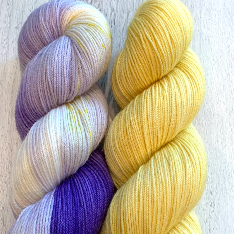 Two skeins of yarn; on the left a white and purple yarn with flecks of yellow, on the right a solid yellow.